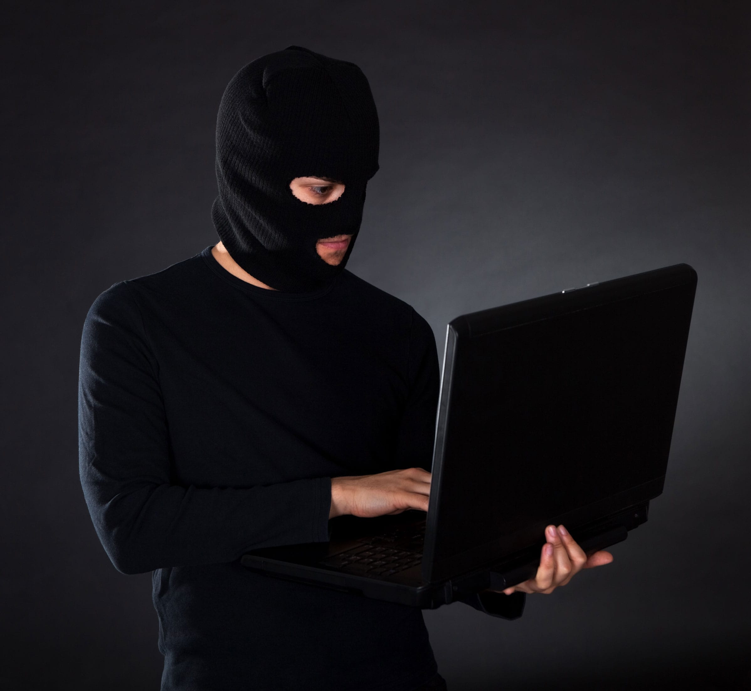 Man wearing a balaclava mask and using a laptop for felony fraud, possibly in violation of California PC 484